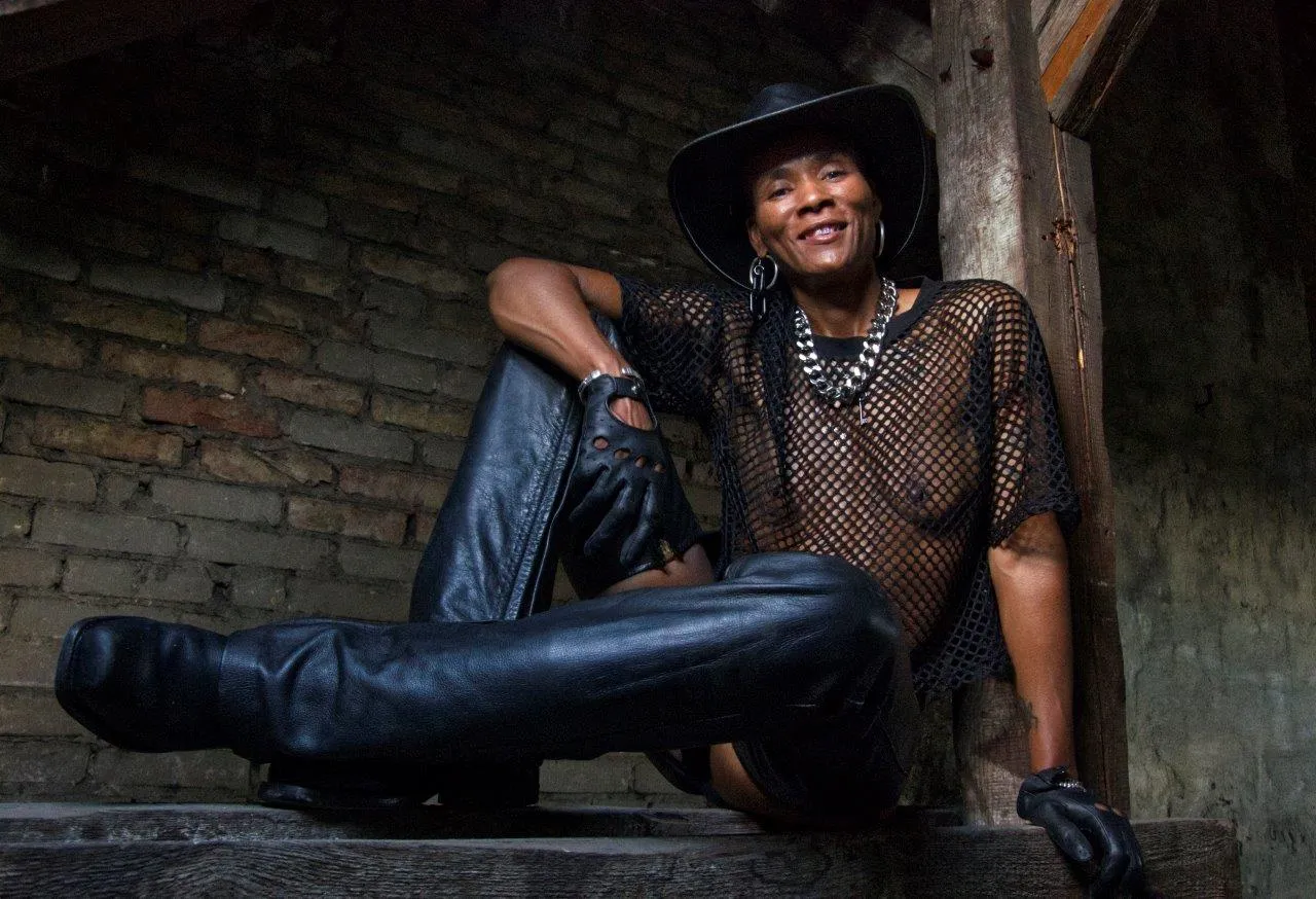 AfroDisiac, a BDSM artist, wearing a leather hat, gloves, chaps, and boots with a mesh top.