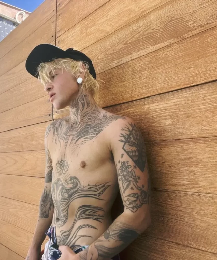 Austin Spears shirtless with tattoos outdoors against a wooden background wearing a black cap.