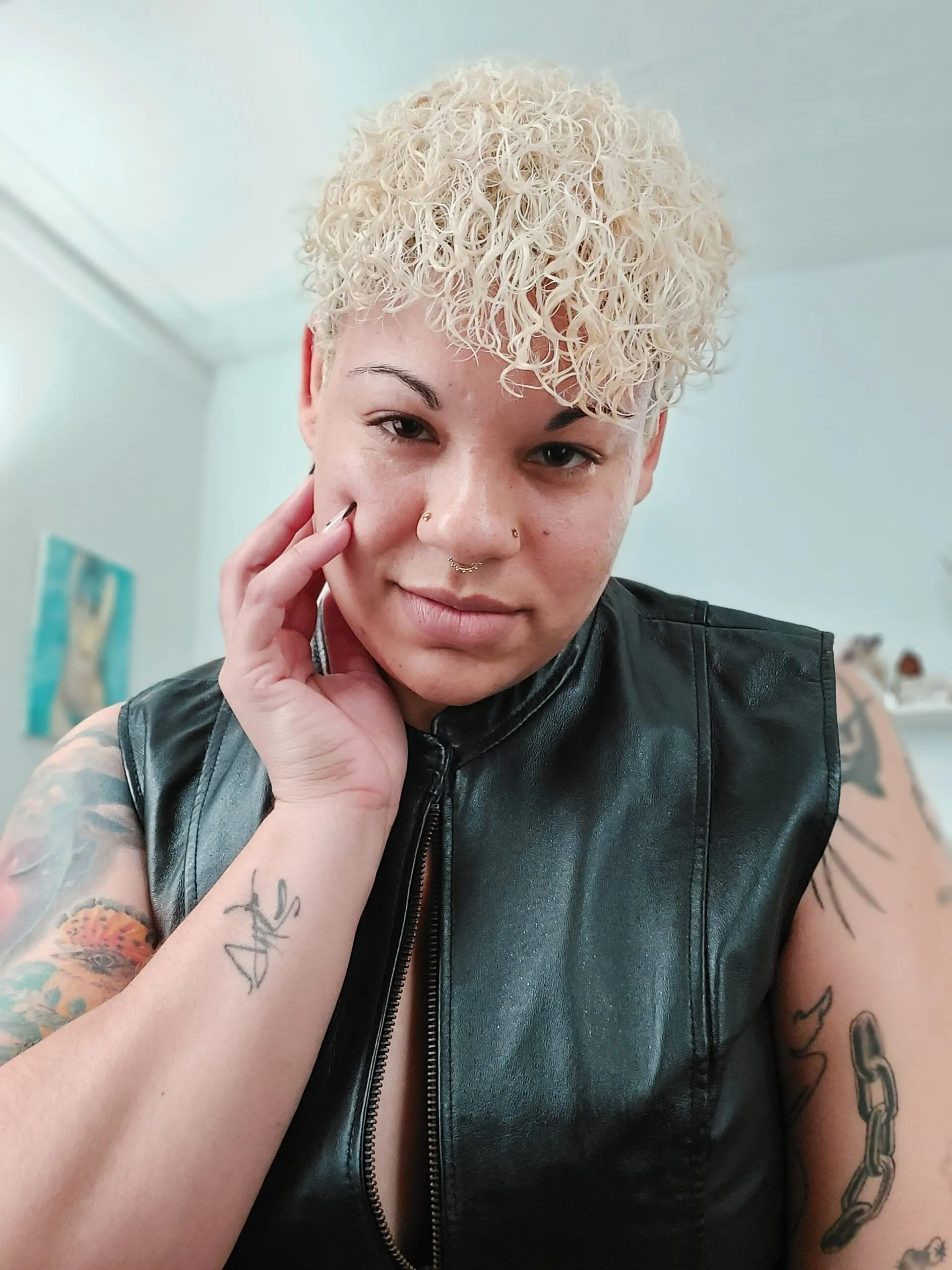 Queer filmmaker, Oran Julius, smirking and staring into the camera as he touches his face while wearing a leather vest.