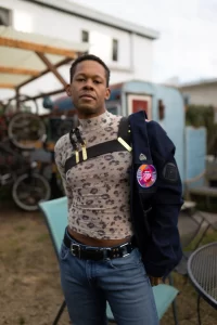 LT Hawk, a black, queer, non-binary pleasure-based sex educator poses confidently outdoors in a floral top, asymettrical black leather chest harness, and jacket slung over their shoulder.