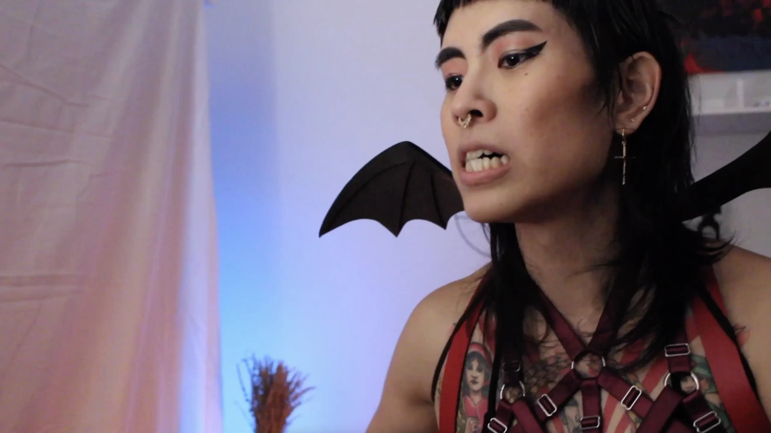 Dahlia Damoiselle, a trans femme erotic performer, bares her teeth while wearing bat wings and cat eye eyeliner.